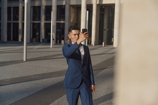 A young asian sales manager in  a blue classic suit makes selfie in the evening light, standing on a square in front of the business center, we see him from the distance with his hand in front of him
