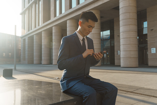 A young asian entrepreneur  in  a blue classic suit and a white shirt is sitting in a stone bench on the square in the evening light he is near the beige business center with columns with his phone in his hands , we see him from aside
