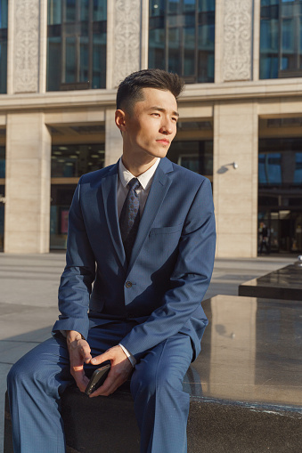 A young asian businessman in  a blue classic suit and a white shirt is sitting in a stone bench on the square near the beige business center with square columns and looks away with his phone in his hands , we see him in the evening light in a profile