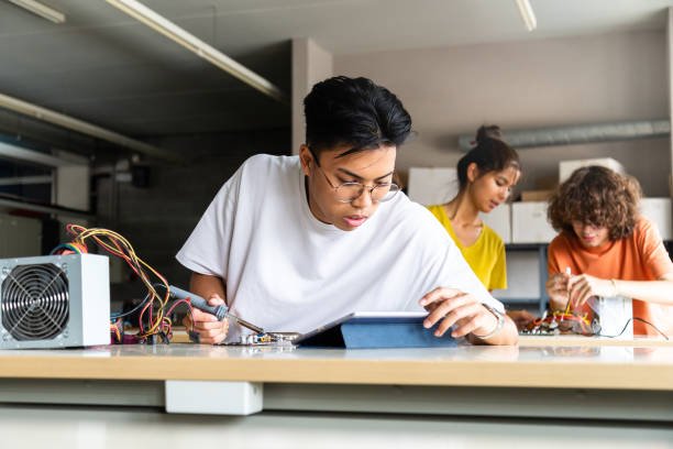 istock Teen asian high school student using tablet to read instructions in electronics class. Classmates in background. 1332657084