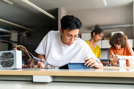 Teen asian high school student using tablet to read instructions in electronics class. Classmates in background. Education concept.