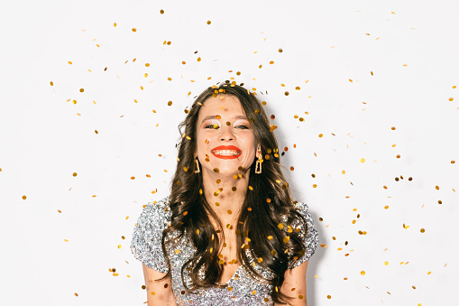 Holiday celebration. Excited woman. Festive decor. Happy beautiful laughing lady enjoying party throwing sparkle golden confetti spangles isolated white.