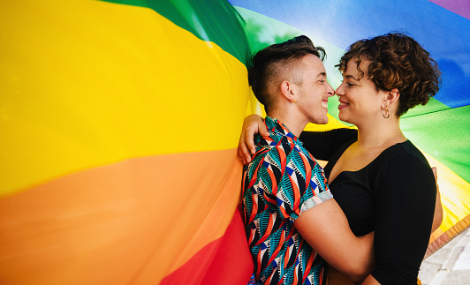 Affectionate queer couple bonding against a rainbow pride flag. Young LGBTQ couple embracing each other and touching their noses together. Two non-conforming lovers smiling while standing together.