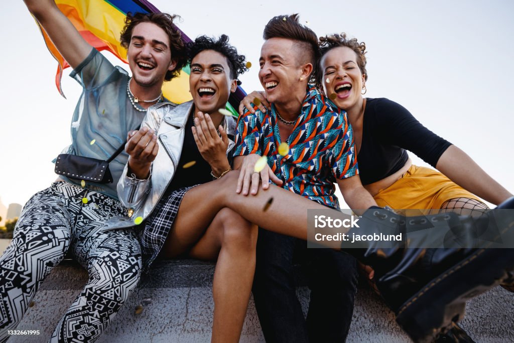 Young people celebrating gay pride outdoors Young people celebrating pride while sitting together. Four members of the LGBTQ+ community smiling cheerfully while raising the pride flag. Group of queer individuals celebrating together outdoors. LGBTQIA Rights Stock Photo