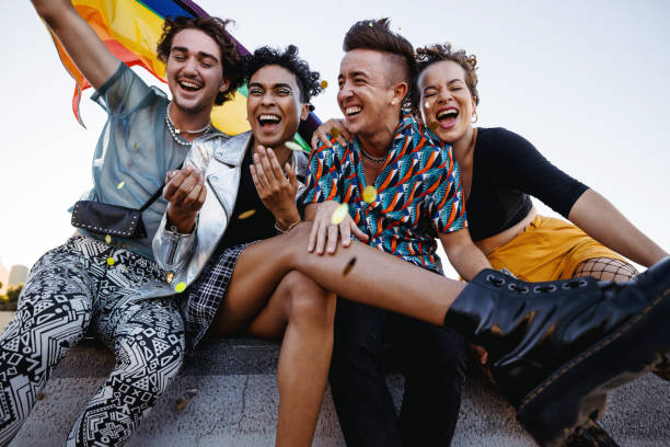 Young people celebrating gay pride outdoors Young people celebrating pride while sitting together. Four members of the LGBTQ+ community smiling cheerfully while raising the pride flag. Group of queer individuals celebrating together outdoors. gay person stock pictures, royalty-free photos & images