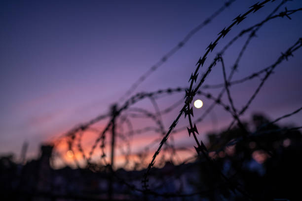 Sunset Behind Wall Barbed wire steel wall against sunset. prison lockdown stock pictures, royalty-free photos & images