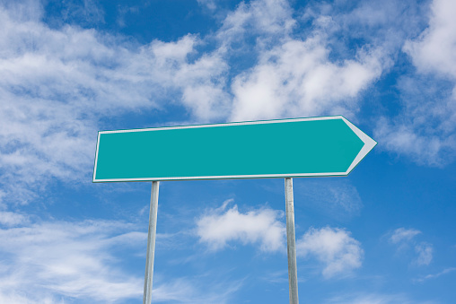 Blank green directional arrow road sign against blue sky with clouds.