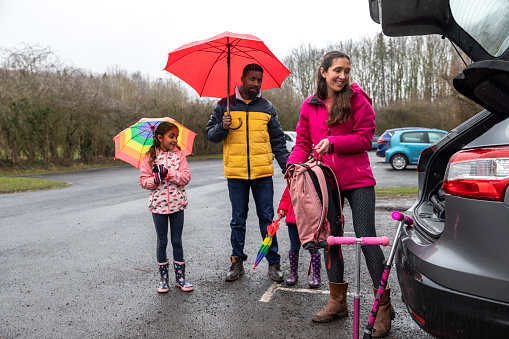 A multi-ethnic family standing in a carpark, getting ready to go on a day trip to Plessey Woods, Northumberland. The father and daughter are holding umbrellas over their heads to protect them from the rain while the mother gets her backpack out of the car boot. There are scooters leaning against the car, ready for the children to use.