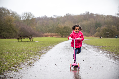A young mixed race girl wearing a padded raincoat, riding a push scooter on a wet footpath through Plessey woods, northumberland. She is smiling while looking ahead.
