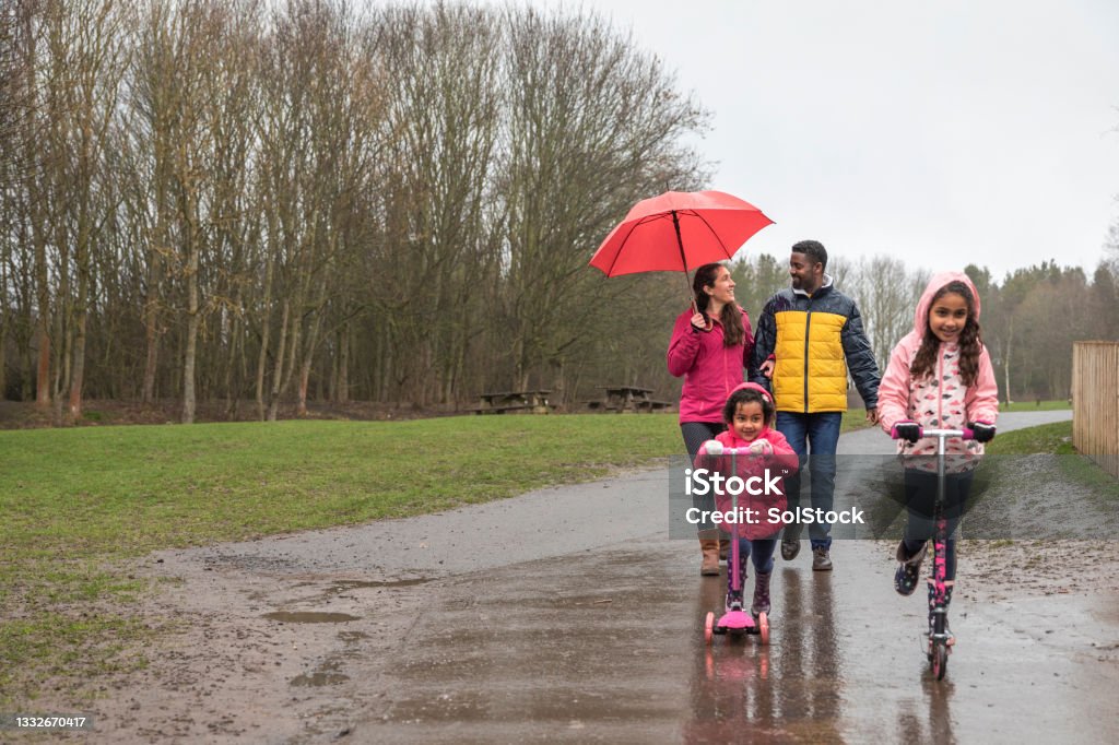 The Perfect Day Out Two young girls riding their push scooters on a wet footpath through Plessey woods, Northumberland. Their parents are walking behind them, the mother is holding an umbrella above her head to shield her from the rain while talking to her husband. They are all enjoying a day out together. Rain Stock Photo
