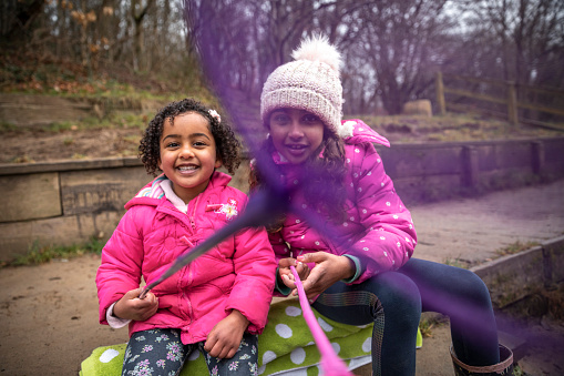 Two young girls wearing padded jackets sitting on a walkway at Plessey Woods, Northumberland. They are holding butterfly fishing nets up to the camera, ready to catch animals in them while looking at the camera and smiling.