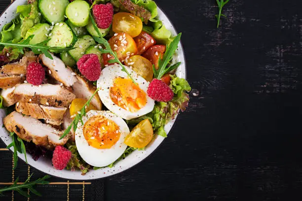 Photo of Grilled chicken meat and fresh vegetable salad of tomato, cucumber, egg, lettuce and raspberry. Ketogenic diet. Buddha bowl dish on dark background. Top view, flat lay