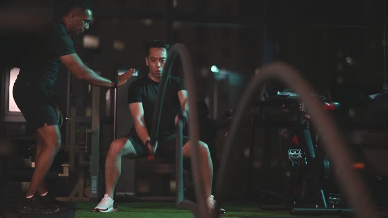 Asian Indian personal trainer encouraging Asian Chinese Mid Adult man practicing challenging battle rope in gym