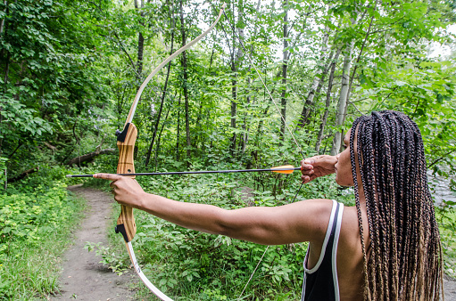 Young Haitian woman aiming at target with bow in forest during day of summer