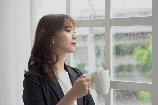 Beautiful Asian businesswoman (Thai, Japanese, Korean or Chinese) with a white coffee cup stands near the window on a rainy day happily and looks out the window.