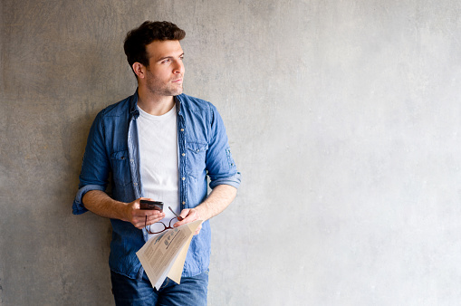 Thoughtful Latin American man using his cell phone and holding documents while leaning against a wall