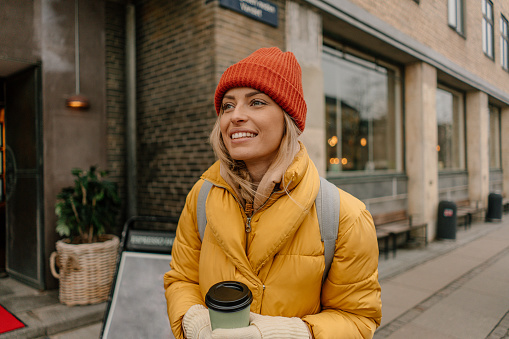 Photo of a smiling young woman finding beauty and joy on a cold winter day in the city; she is sipping coffee-to-go while walking the city streets