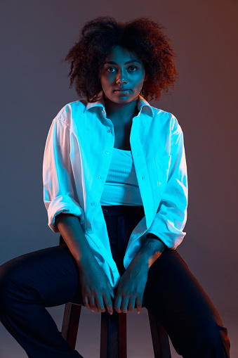 Calm and beautiful. Full-length portrait of African young girl casual clothes, outfit isolated on dark blue studio background in neon light. Concept of human emotions, facial expression, youth, ad.