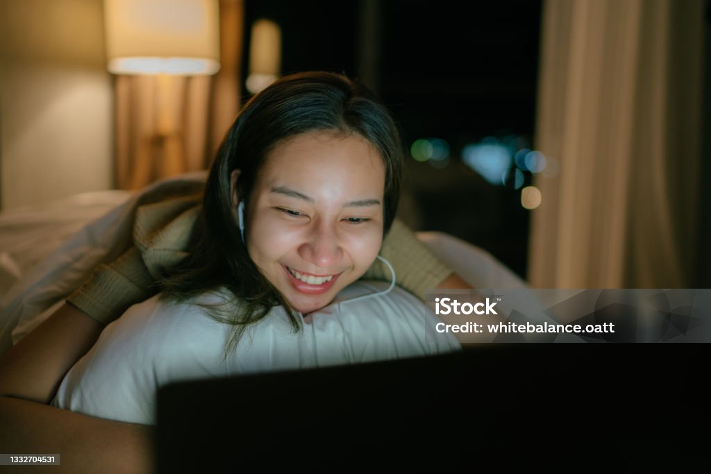 Asian woman relaxing on Friday night at her apartment watching a movie on TV enjoying a night in bed. Asian woman relaxing on Friday night at her apartment watching a movie on laptop enjoying a night in bed. Movie Stock Photo
