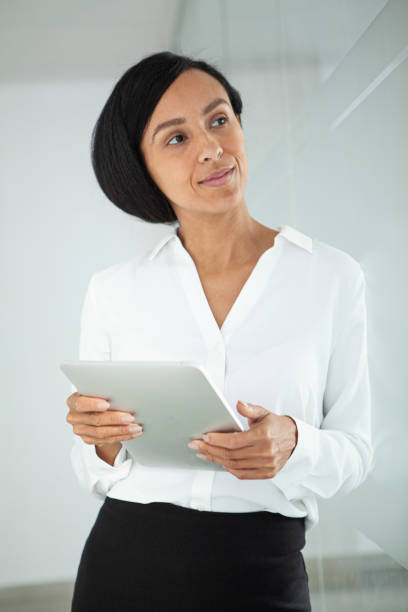 Medium shot of thoughtful latin businesswoman looking up while holding digital tablet standing in corridor during daytime