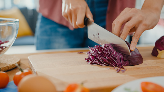 Hand of young Asian woman chef hold knife cutting Red Chinese cabbage on wooden board on kitchen table in house. Cooking vegetable salad, Lifestyle healthy food eating and traditional natural concept.