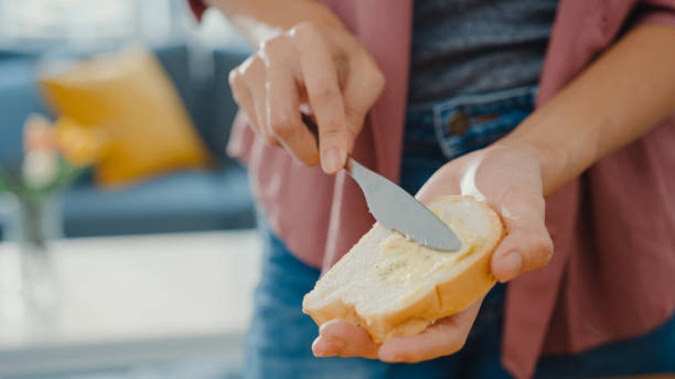 hände einer jungen asiatischen köchin, die butter auf rustikalem roggenbrot mit metallmesser auf holzbrett auf küchentisch im haus verteilt. - butter margarine fat bread stock-fotos und bilder