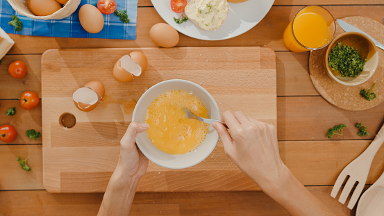 Hands of young Asian woman chef whisking egg into ceramic bowl cook omelette with vegetables on wooden board on kitchen table in house. Lifestyle healthy eat and traditional bakery. Top view shot.
