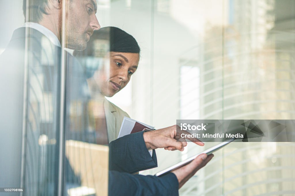 Successful latin businessman standing behind window using digital tablet with businesswoman Medium side shot of successful latin businessman standing behind window using digital tablet to discuss information with businesswoman while standing in office during day Lead Stock Photo