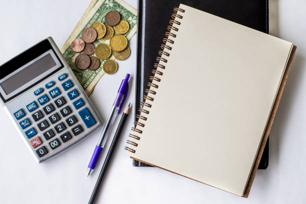 istock Plain Spiral Notebook With Pen Beside Calculator And Coins On Table. Simple Notepad And Ballpen Alongside Calculation Machine Placed On Top Of Desk. 1332712839