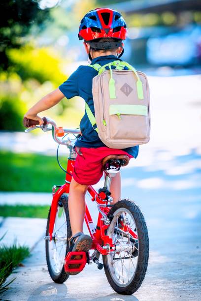 little boy riding his bicycle going back to school carrying his backpack wearing a bicycle helmet from behind - little boys preschooler back to school backpack imagens e fotografias de stock