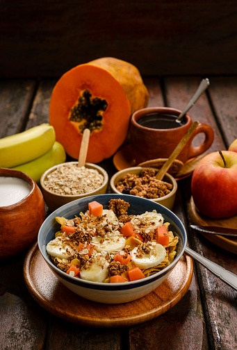 Rustic wood table with healthy food ingredients at home in the kitchen with fruits as banana, papaya, red apple, oat flake, crunch flake and granola cereal. Healthy lifestyle living isolated from people and pandemic times of Covid-19 and variants as the Delta.Image made in studio with a 24 megapixels camera with full frame size sensor and 55 mm macro lens f/stop 2.8