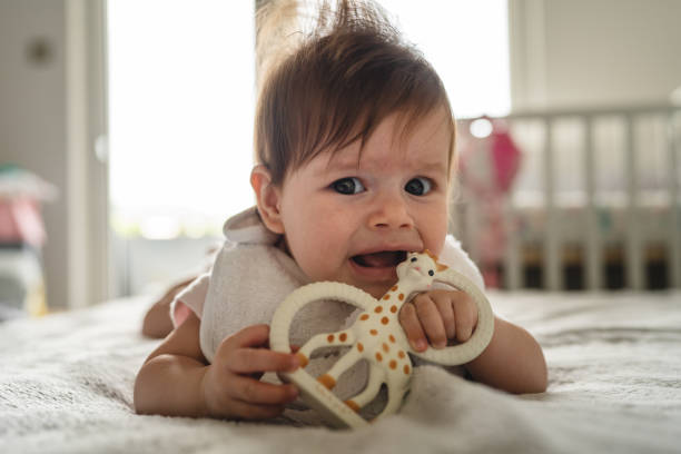 Small caucasian baby four or five months old having teeth growing issues teething pain while holding a bite toy looking to the camera lying on the bed with teether at home