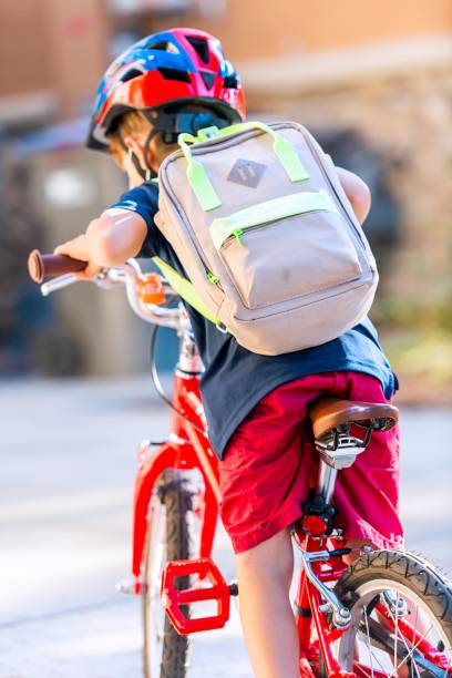 istock Little boy riding his bicycle going back to school carrying his backpack wearing a bicycle helmet from behind 1332739256