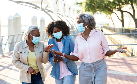 A group of three senior African-American women walking side by side outdoors, on a city waterfront, having an animated conversation. They are wearing protective face masks, trying to prevent the spread of coronavirus during the covid-19 pandemic. They are smiling and laughing behind their masks.