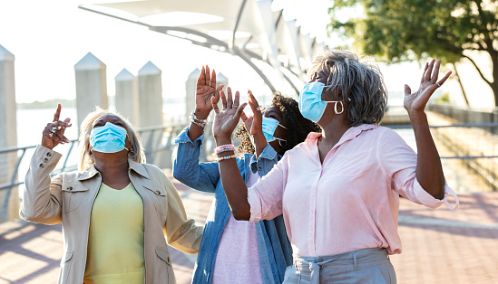 A group of three senior African-American women standing together outdoors, on a city waterfront, looking up with their hands raised. They are wearing protective face masks, trying to prevent the spread of coronavirus during the covid-19 pandemic. They are smiling and laughing behind their masks.