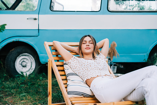 Young woman resting in nature, sitting on wooden chair near van outdoors. Enjoying summer, travel concept