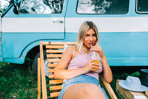 Portrait of young woman sitting on wooden chair near van and drinking lemonade outdoors in nature. Enjoying summer, travel concept