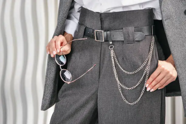 Gray women's business trousers, with a high waist and a chain on the belt. A fragment of the model's body in a jacket, trousers and a white shirt. A woman holds round-rimmed glasses in her hand.