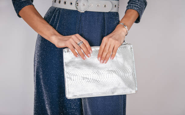 Women's silver clutch bag with snakeskin texture. Women's fashion accessories.Graceful hands in rings and bracelets hold a silver clutch. A woman in a blue sequined dress with a wide gray belt. older woman purse stock pictures, royalty-free photos & images
