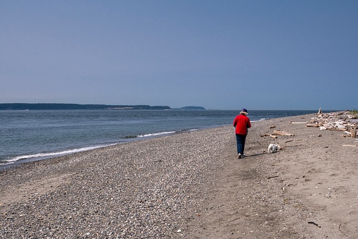 A senior woman, dressed in a red jacket, walking her white dog on a beach on Whidbey Island which is located in Puget Sound, in the state of Washington.