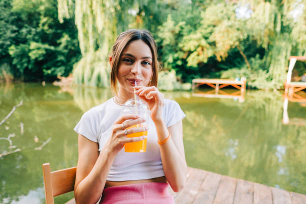 młoda atrakcyjna kobieta pijąca pomarańczową lemoniadę, odpoczywająca na molo nad jeziorem na świeżym powietrzu... - juice drinking women drink zdjęcia i obrazy z banku zdjęć