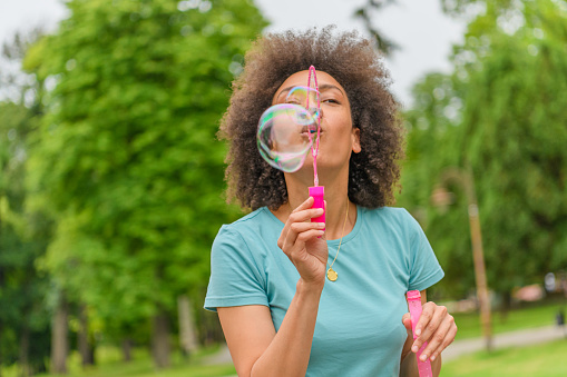 Young Mother is Walking Through the Park and Having a Fun Blowing Bubbles.