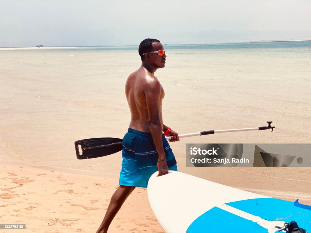 Young man taking paddle board int owater. Young man walking on the beach with paddle board and paddle in his hands. Golden and smooth sand on Egypt seashore. Men Stock Photo