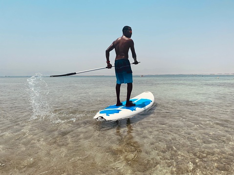Young man in swimming shorts is standing on the board, paddling, away from the seashore, enjoying in fresh sea breeze, blue sky and clear and calm water.