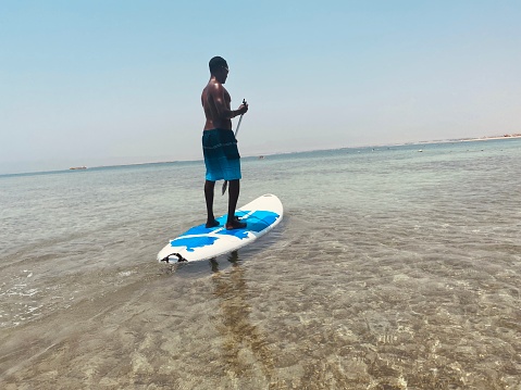 Attractive man in swimwear floating on stand up paddle board on a quiet blue sea. Surfing in tropical sea. Beautiful day on Egypt beach.