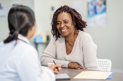 A middle aged woman smiles as she discusses treatment options with her doctor in an office. There are forms on the table in front of them.