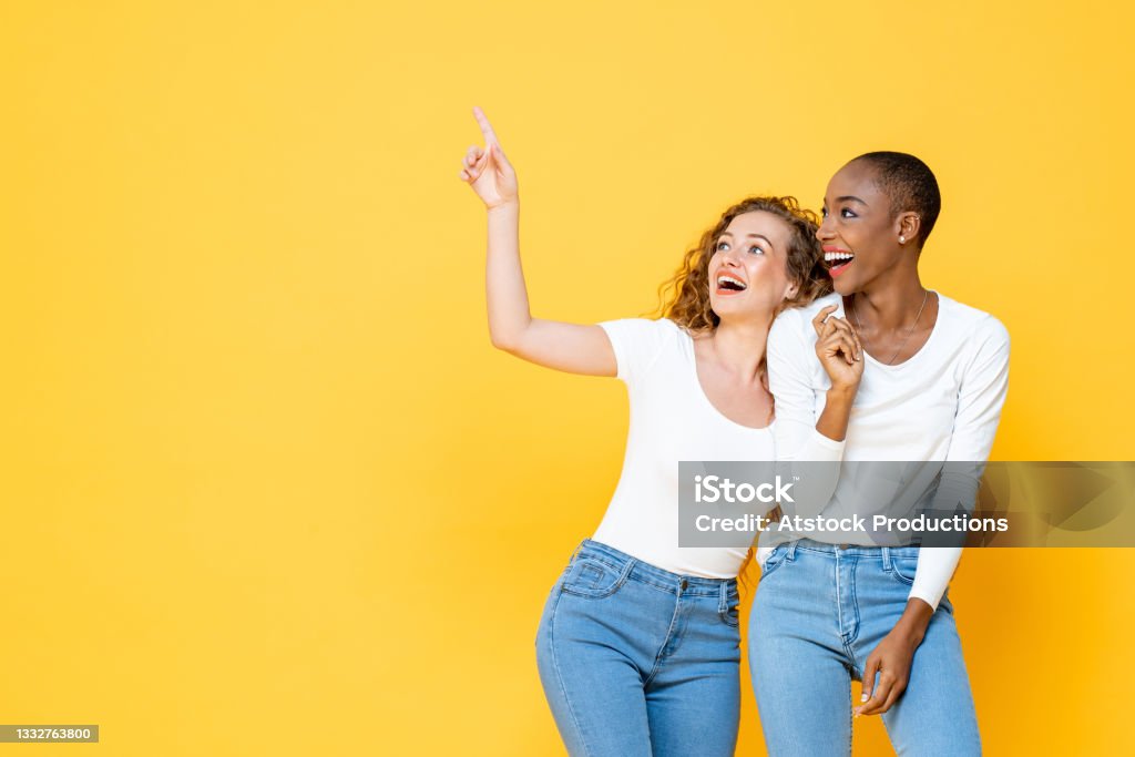 Joyful happy interracial woman friends pointing and looking up to empty space on isolated yellow studio background Friendship Stock Photo