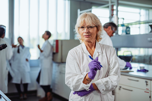 Portrait of blonde woman in late 50s wearing eyeglasses, lab coat, purple gloves, and looking at camera with arms crossed while standing in educational lab.