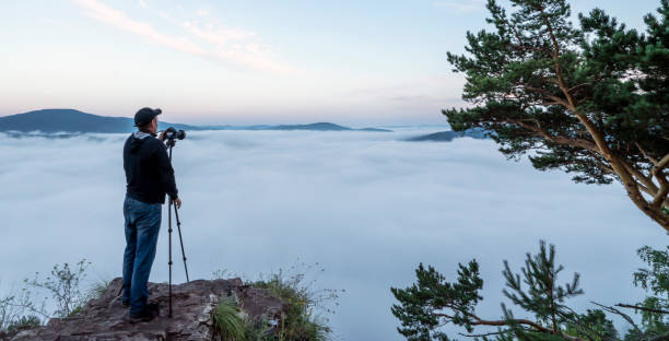 a man photographer in the mountains takes pictures with a camera on a tripod of nature and the morning fog of the hills. the early time is dawn and mist. landscape artist and travel photo. love of traveling and passion for the mountains, hiking. un hombre fotógrafo en las montañas toma fotos con una cámara en un trípode de la naturaleza y la niebla de la mañana de las colinas. el tiempo temprano es el amanecer y la niebla. paisajista y foto de viaje. amor por viajar y pasión por las montaña