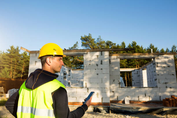 istock Construction worker talks on a smartphone in a yellow hardhat and reflective vest against the background of the construction of house-the walls and window openings are made of porous concrete blocks. 1332793704